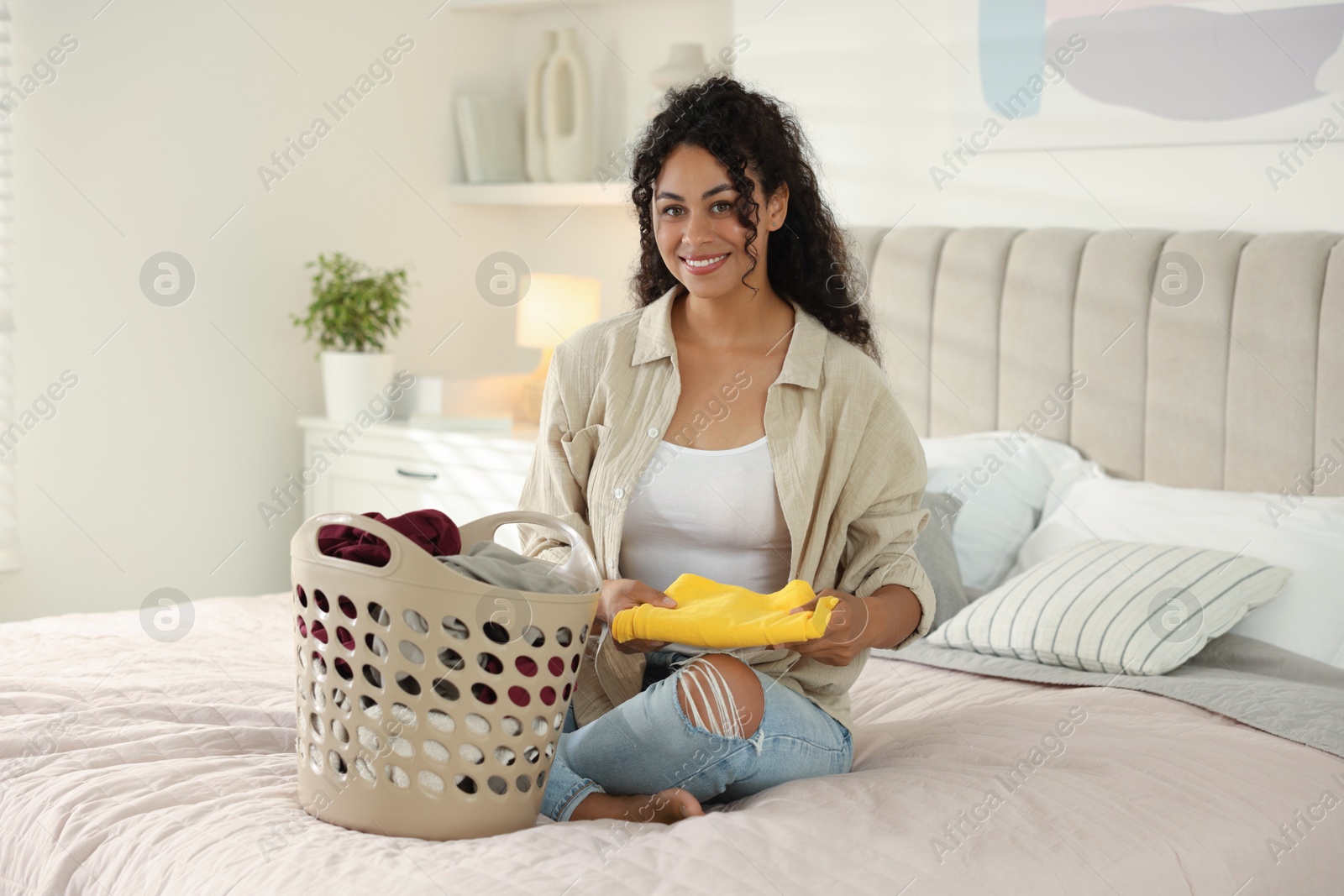 Photo of Happy woman with basket full of laundry on bed at home