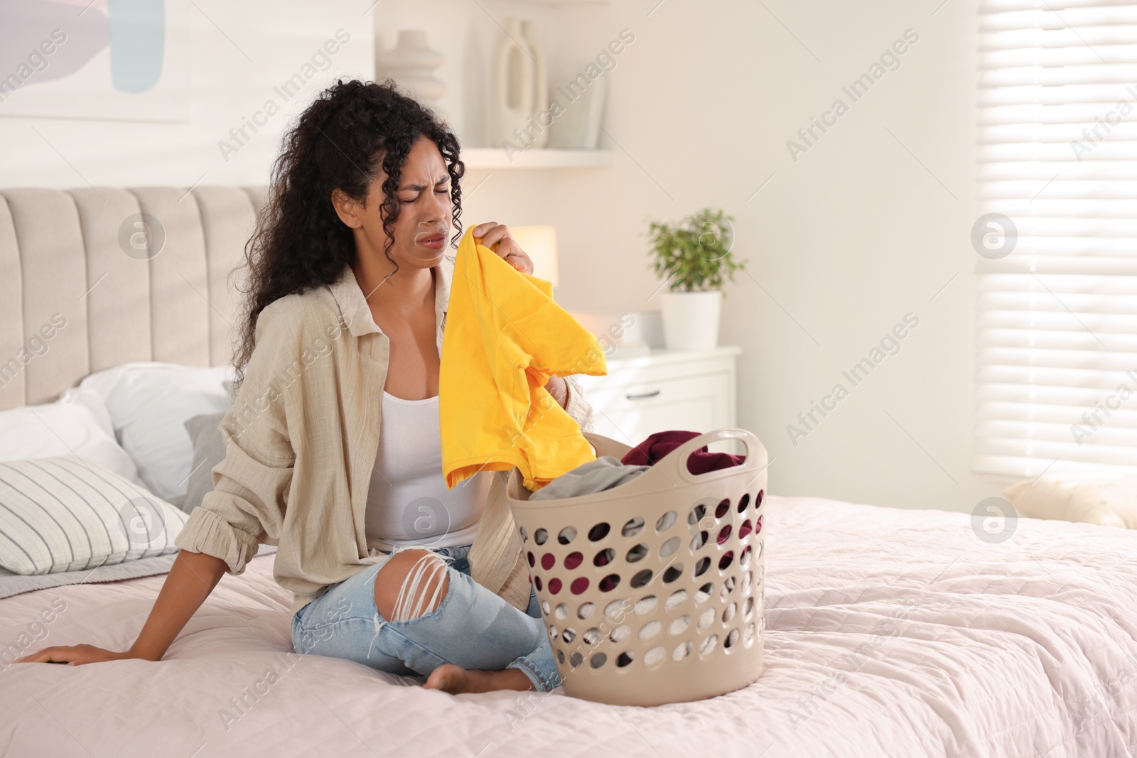 Photo of Displeased woman with basket full of laundry on bed at home, space for text