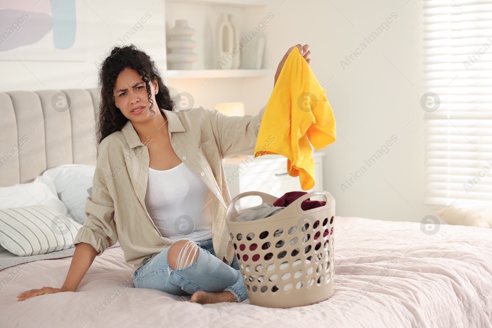 Photo of Displeased woman with basket full of laundry on bed at home, space for text