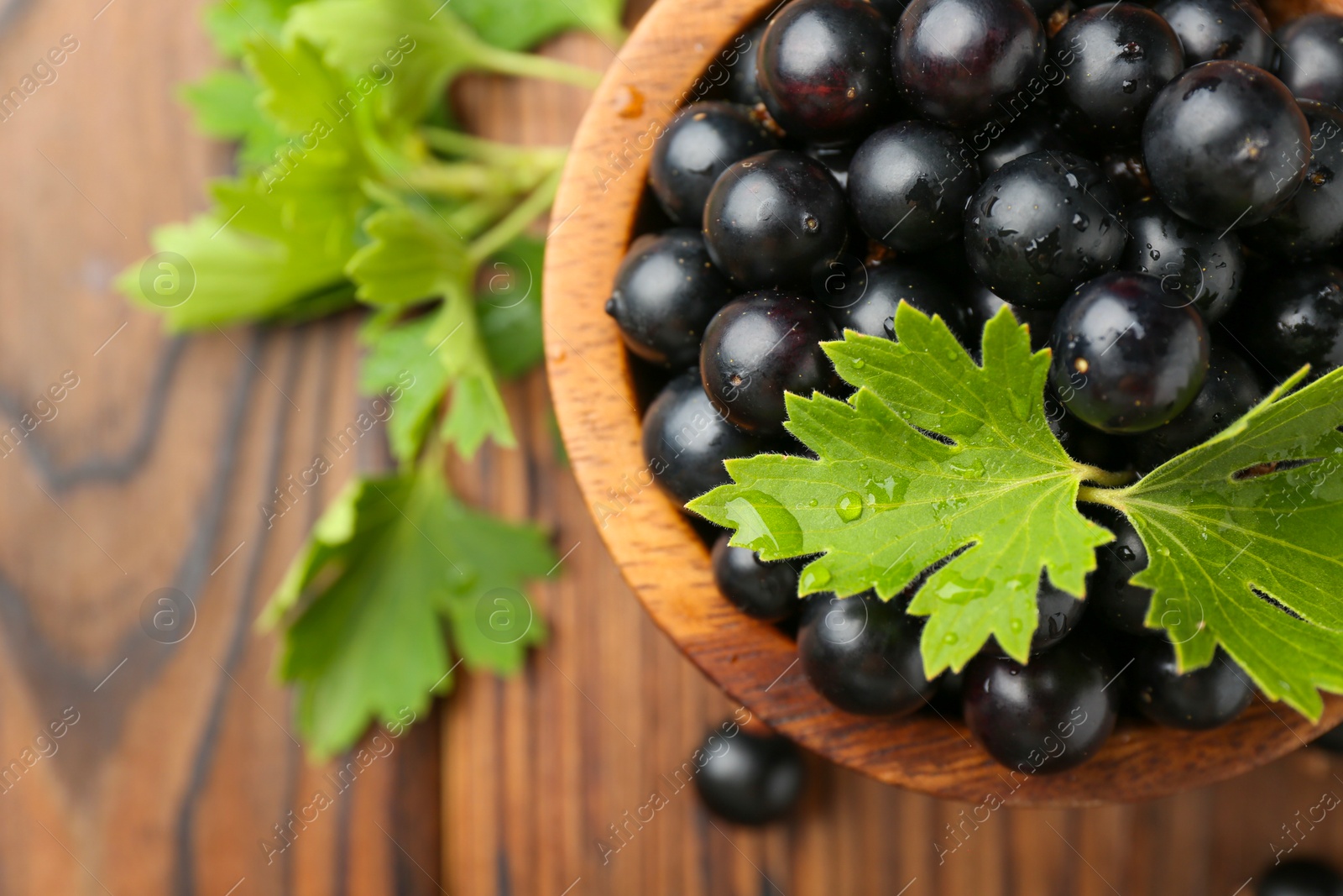 Photo of Ripe black currants and leaves in bowl on wooden table, top view. Space for text