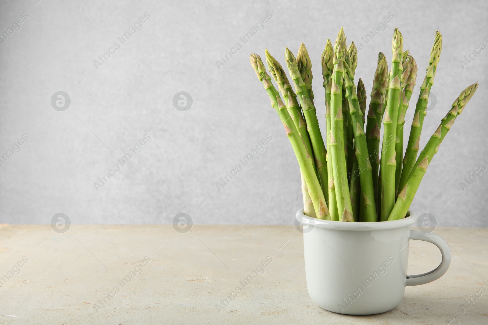 Photo of Fresh green asparagus stems in cup on gray table, space for text