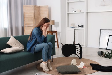 Desperate woman sitting on sofa in messy living room after robbery