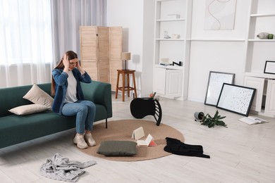 Desperate woman sitting on sofa in messy living room after robbery