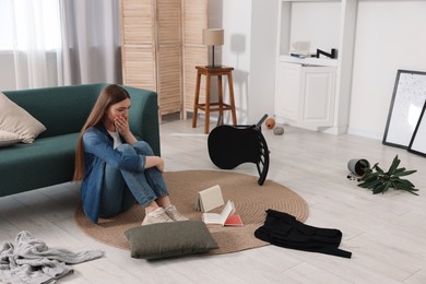 Photo of Desperate woman sitting on floor in messy living room after robbery