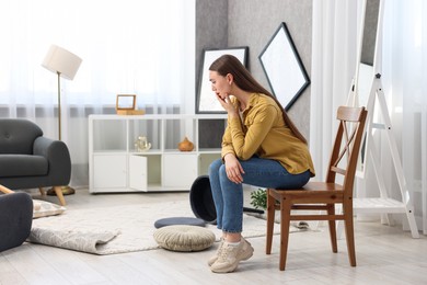 Desperate woman sitting on chair in messy living room after robbery