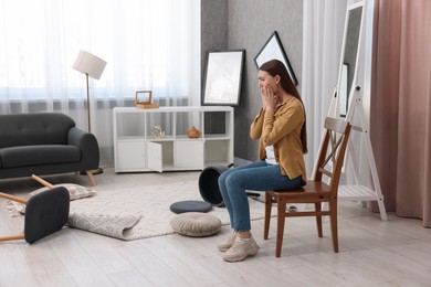 Desperate woman sitting on chair in messy living room after robbery