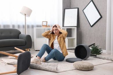 Photo of Desperate woman sitting on floor in messy living room after robbery