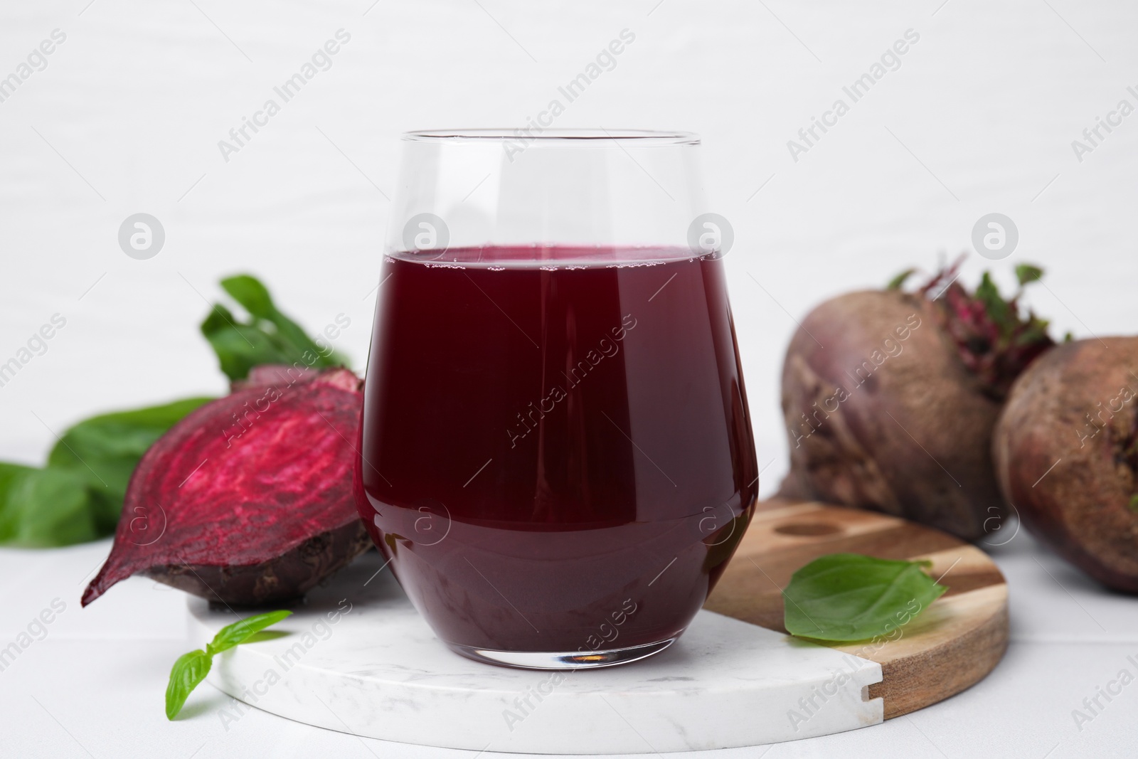 Photo of Fresh beet juice in glass, ripe vegetables and basil on white table, closeup