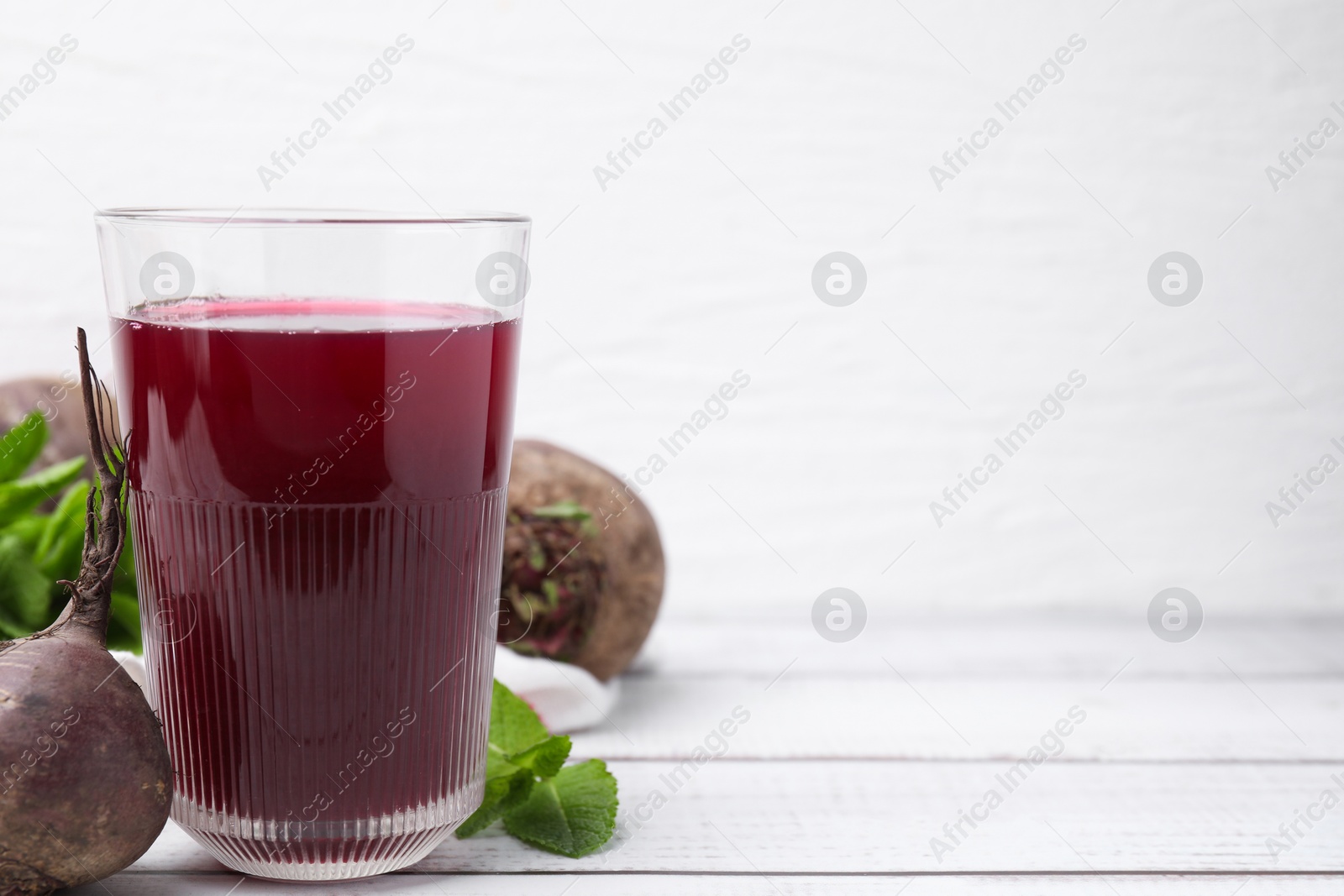 Photo of Fresh beet juice in glass, ripe vegetables and mint on white wooden table, space for text