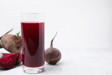 Fresh beet juice in glass and ripe vegetables on white tiled table, space for text