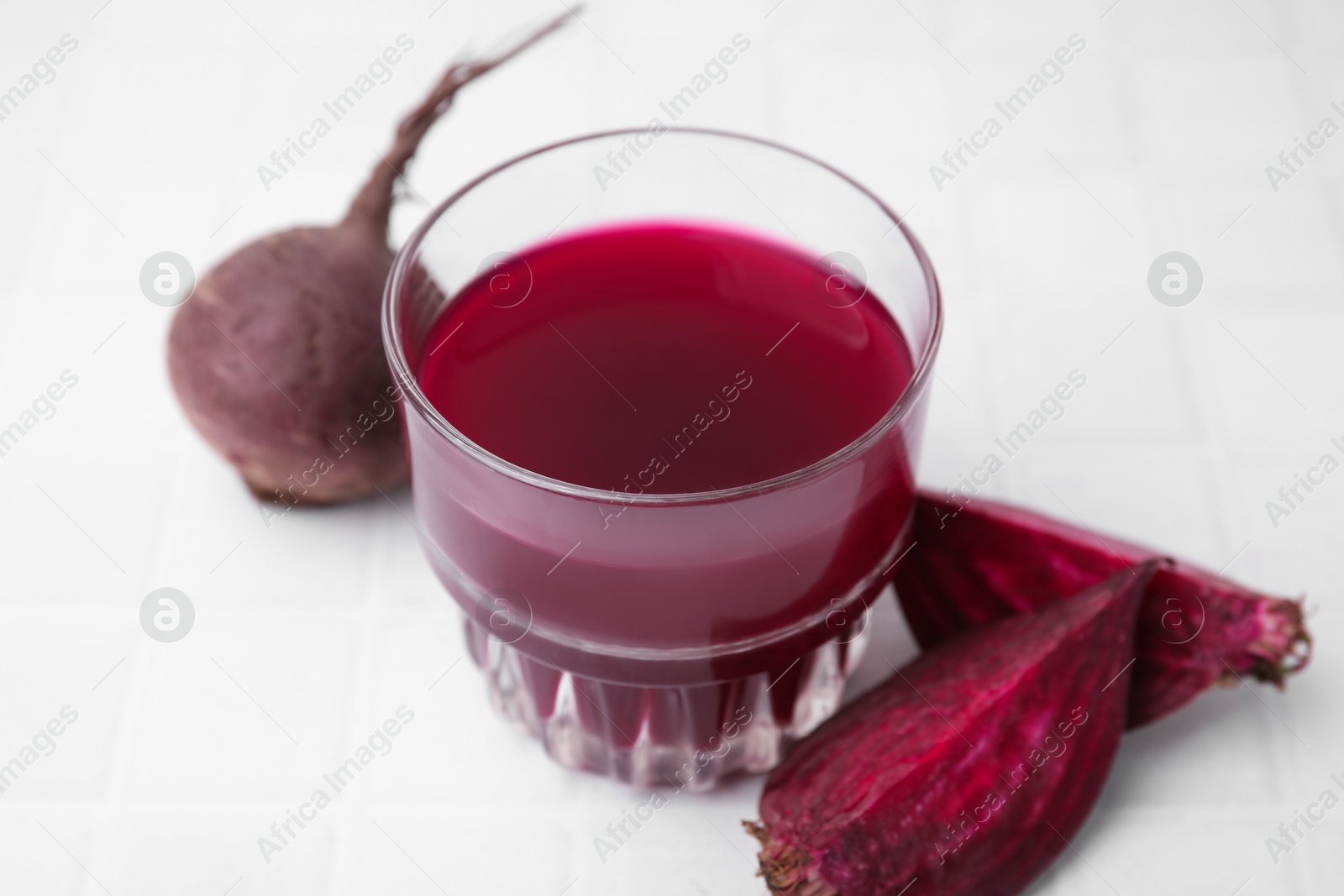 Photo of Fresh beet juice in glass and ripe vegetables on white tiled table, closeup