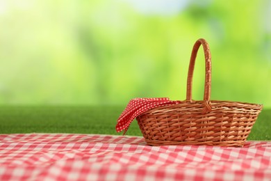 Photo of One picnic wicker basket with checkered napkin and blanket on grass against blurred green background. Space for text