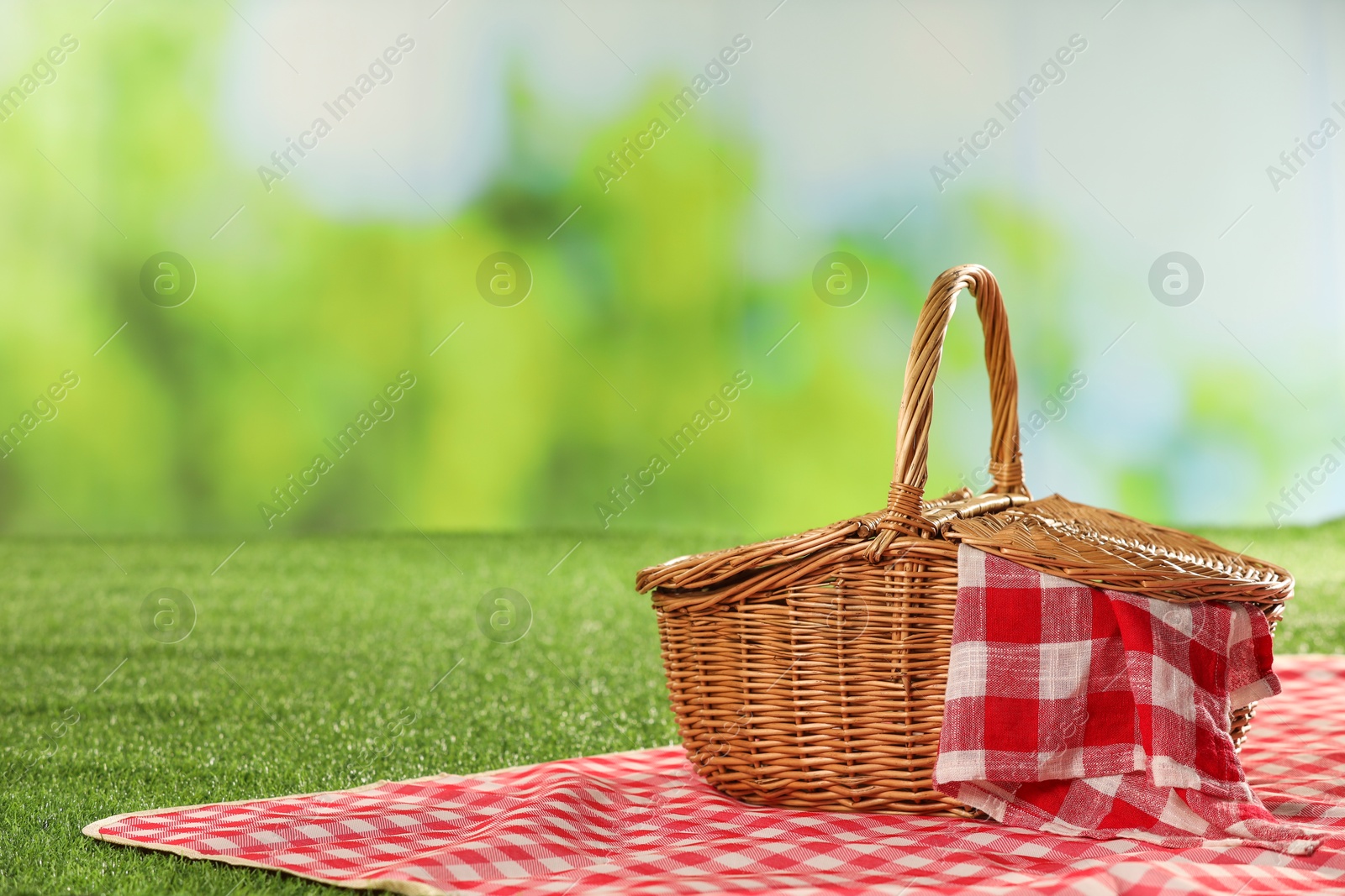 Photo of One picnic wicker basket with checkered napkin and blanket on grass against blurred green background. Space for text