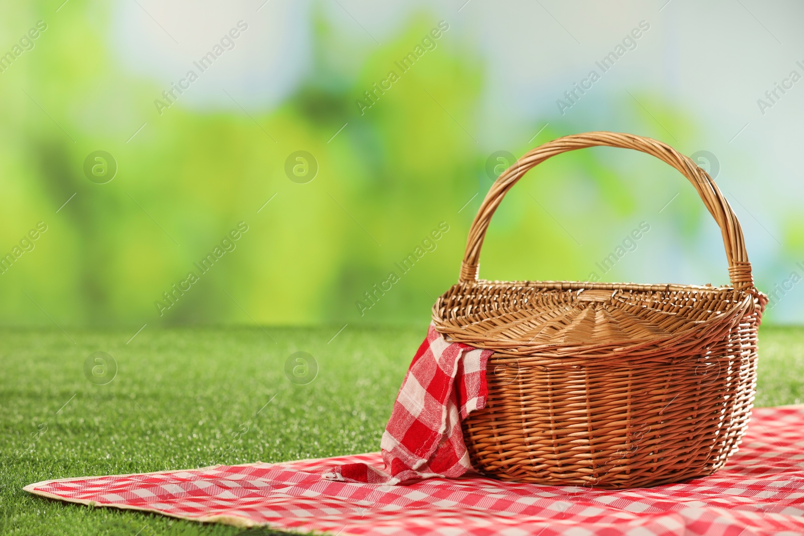 Photo of One picnic wicker basket with checkered napkin and blanket on grass against blurred green background. Space for text