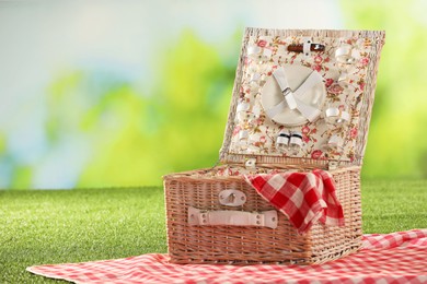 Photo of One picnic wicker basket with checkered napkin, tableware and blanket on grass against blurred green background. Space for text