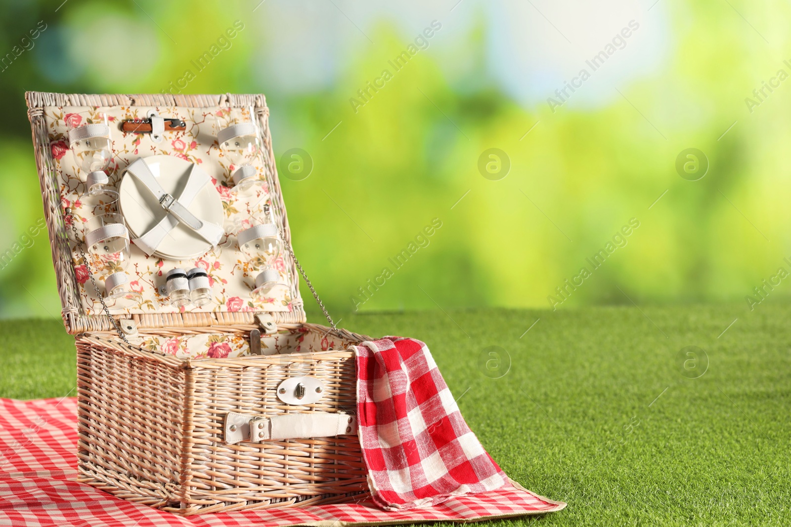 Photo of One picnic wicker basket with checkered napkin, tableware and blanket on grass against blurred green background. Space for text