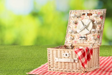 Photo of One picnic wicker basket with checkered napkin, tableware and blanket on grass against blurred green background. Space for text