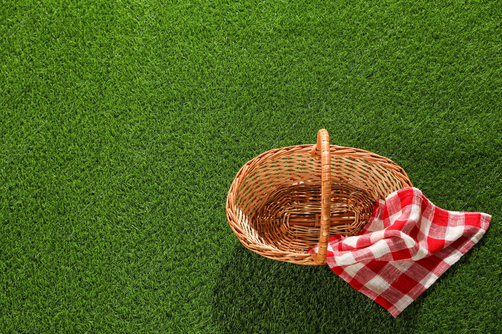 Photo of One picnic wicker basket with checkered napkin on green grass, top view. Space for text