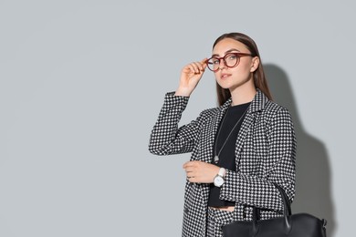 Beautiful woman in stylish suit and glasses with bag on gray background, space for text