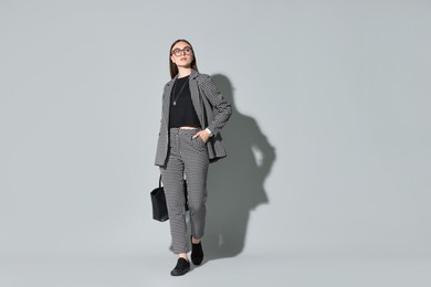 Beautiful woman in stylish suit and glasses with bag on gray background