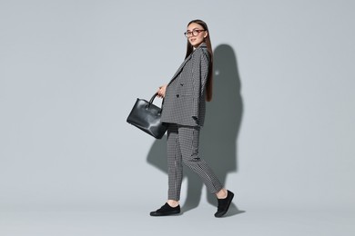 Beautiful woman in stylish suit and glasses with bag on gray background