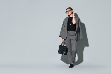 Beautiful woman in stylish suit and glasses with bag on gray background