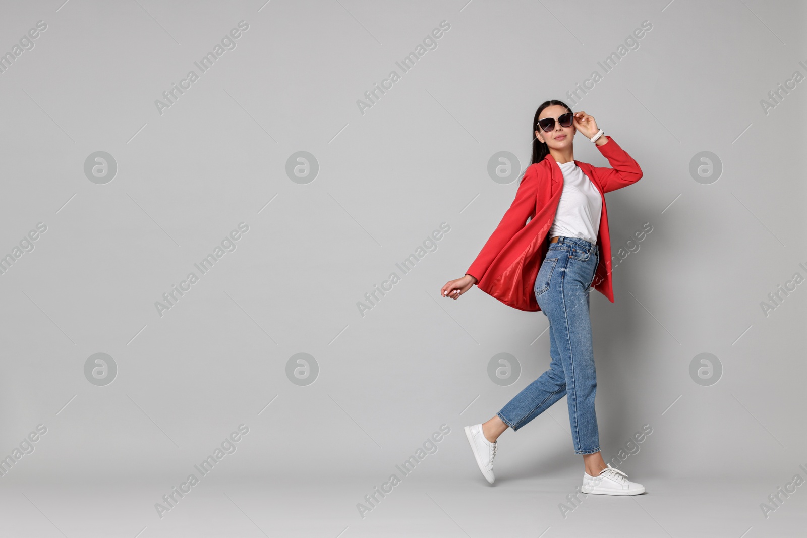 Photo of Stylish woman in red jacket, jeans and sunglasses walking on gray background, space for text