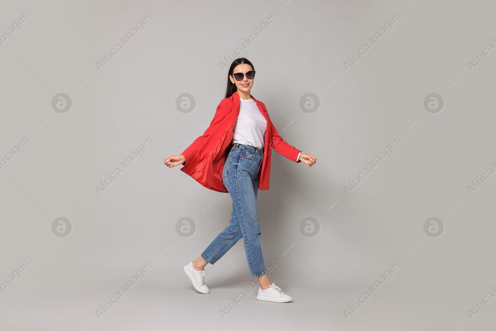 Photo of Stylish woman in red jacket, jeans and sunglasses walking on gray background