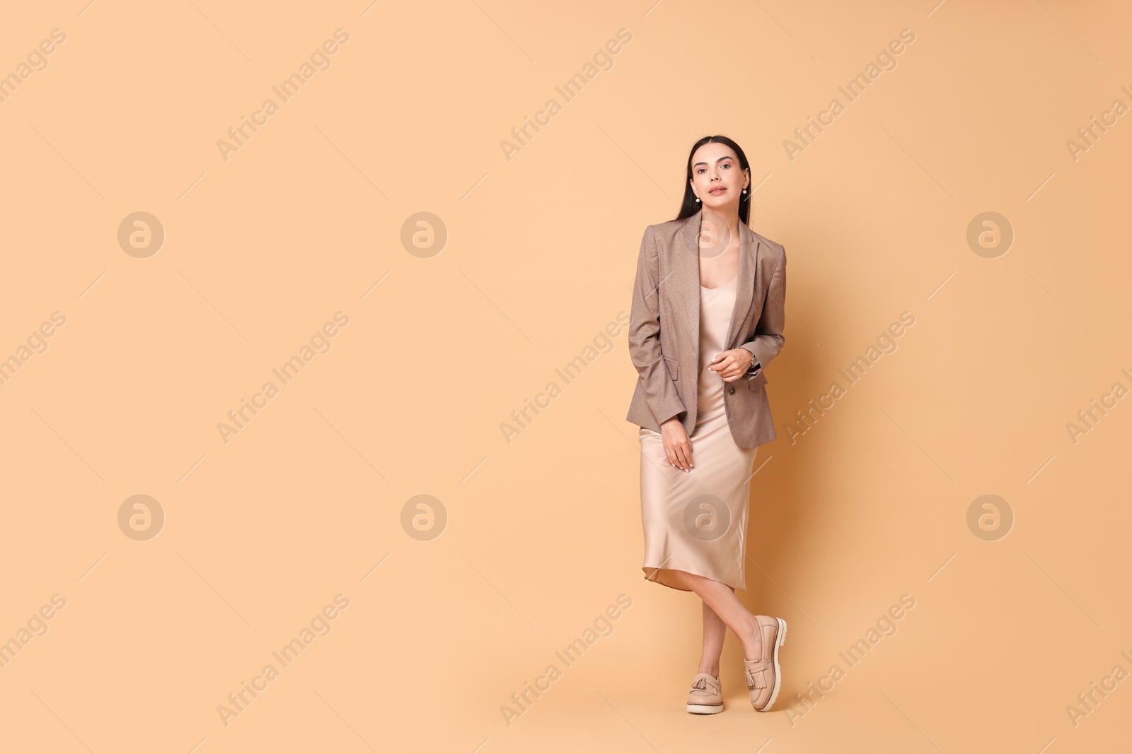 Photo of Beautiful woman in stylish jacket and beige dress on pale orange background, space for text