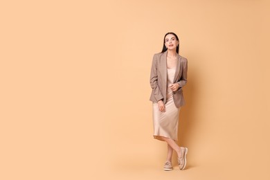 Photo of Beautiful woman in stylish jacket and beige dress on pale orange background, space for text