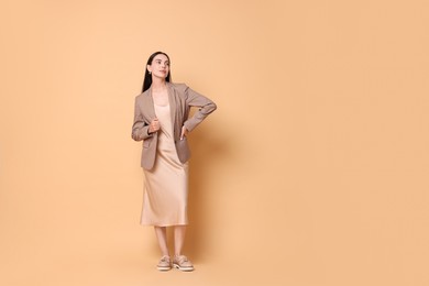 Beautiful woman in stylish jacket and beige dress on pale orange background, space for text