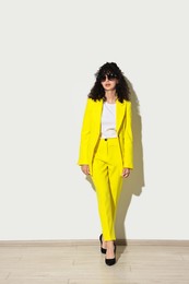 Beautiful young woman in stylish yellow suit and sunglasses near light wall