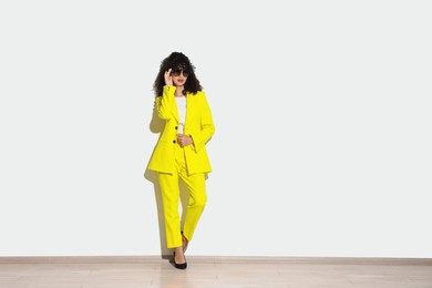 Beautiful young woman in stylish yellow suit and sunglasses near light wall, space for text