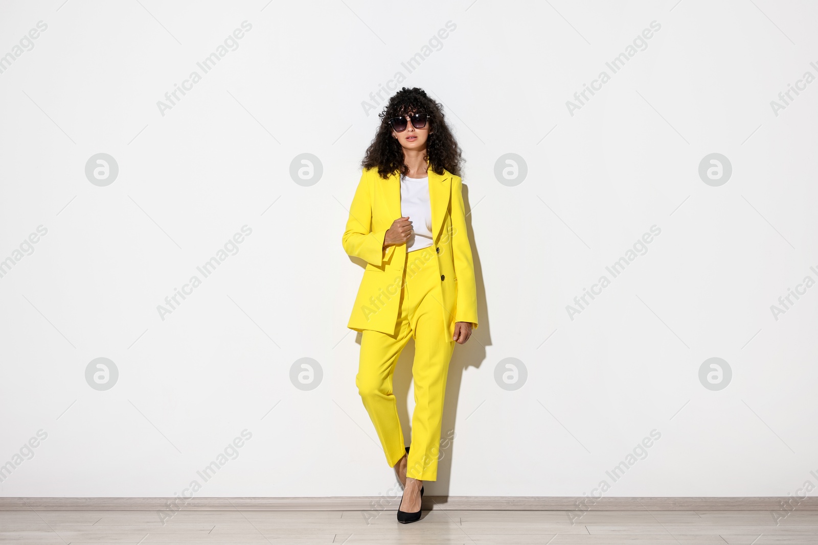 Photo of Beautiful young woman in stylish yellow suit and sunglasses near light wall