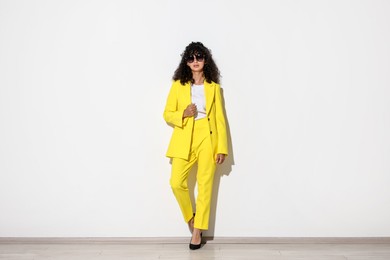 Beautiful young woman in stylish yellow suit and sunglasses near light wall