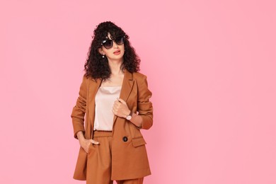 Beautiful young woman in stylish suit and sunglasses on pink background, space for text