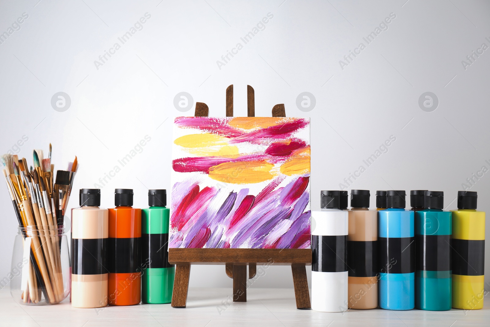 Photo of Acrylic paints of different colors, small easel with abstract picture and brushes on wooden table