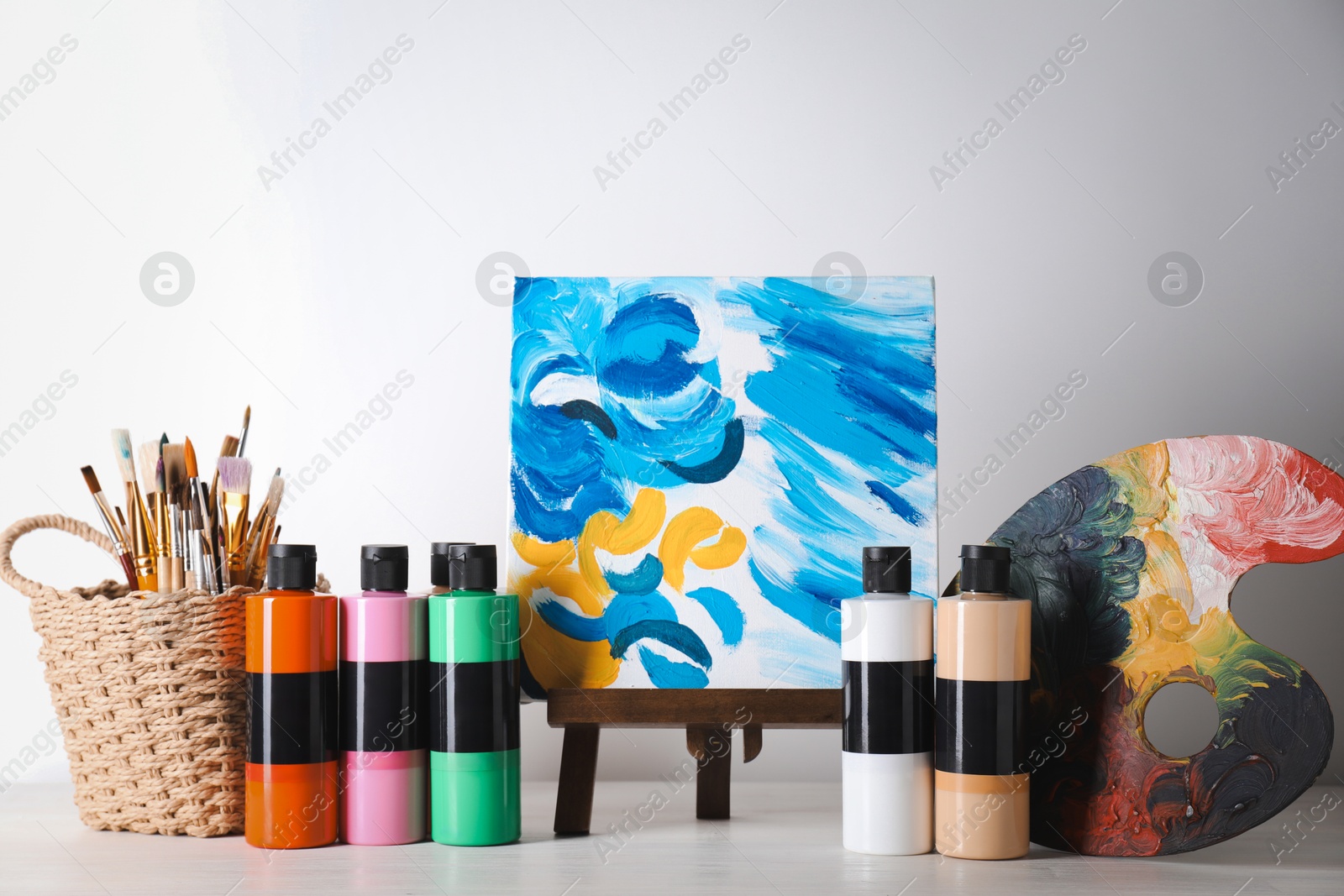 Photo of Acrylic paints of different colors, small easel with abstract picture, palette and brushes on wooden table