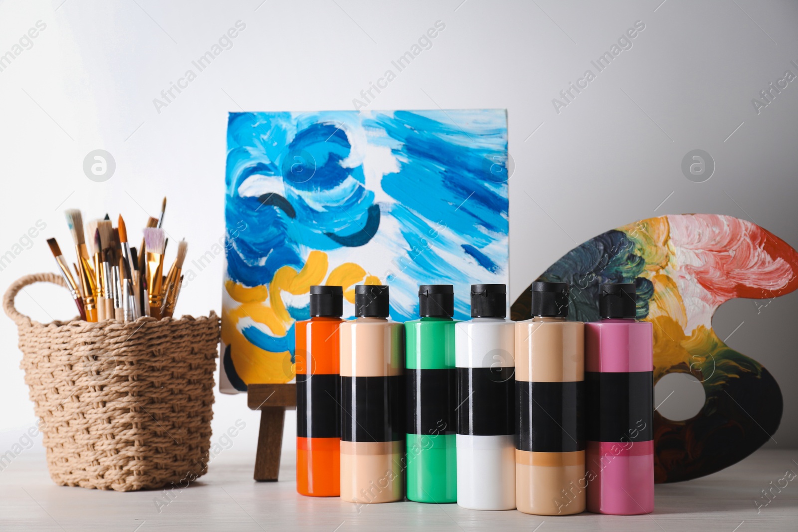 Photo of Acrylic paints of different colors, small easel with abstract picture, palette and brushes on wooden table