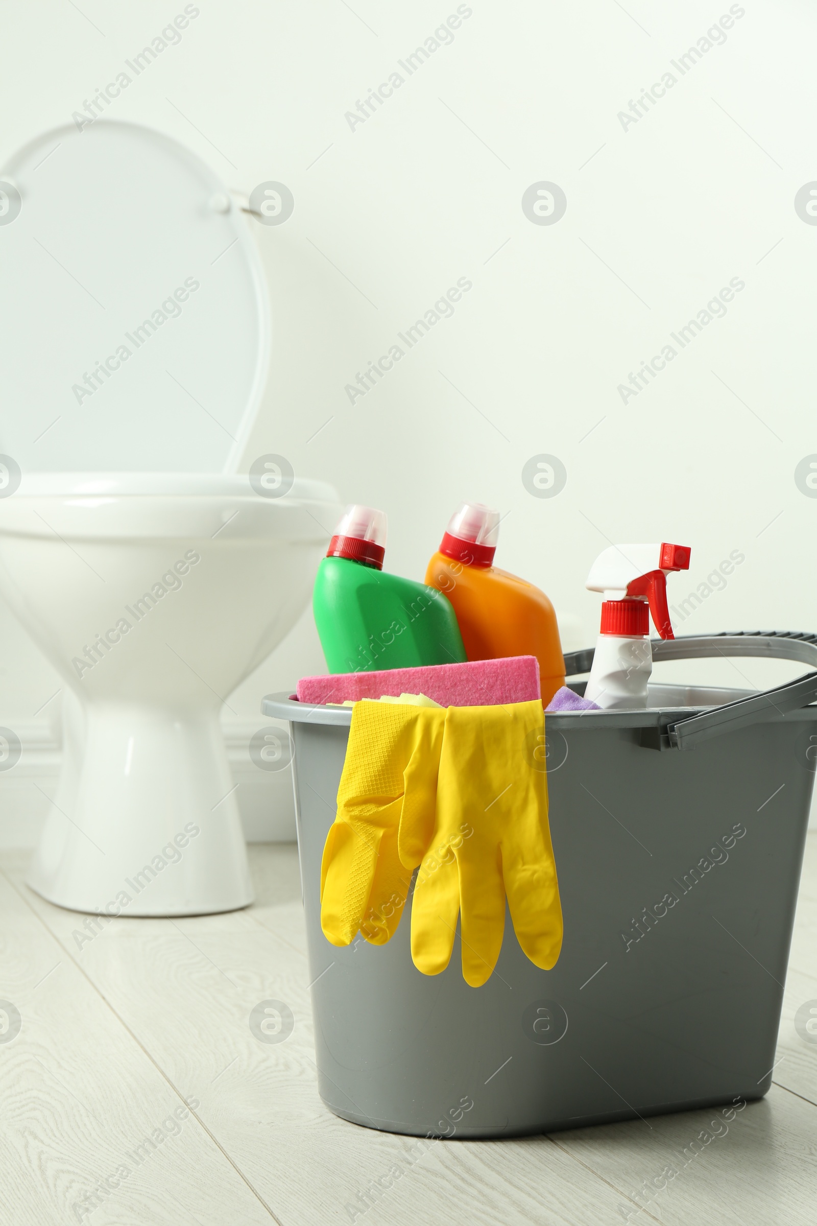 Photo of Bucket with different toilet cleaners, rag and gloves on floor in bathroom