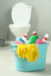 Photo of Bucket with different toilet cleaners, rag, sponge and gloves on floor in bathroom