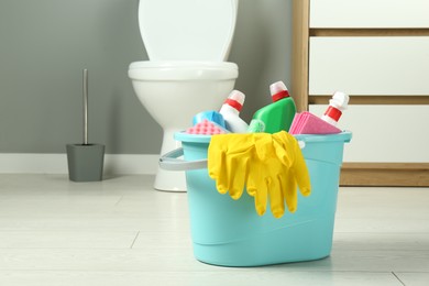 Photo of Bucket with different toilet cleaners, rag, sponge and gloves on floor in bathroom