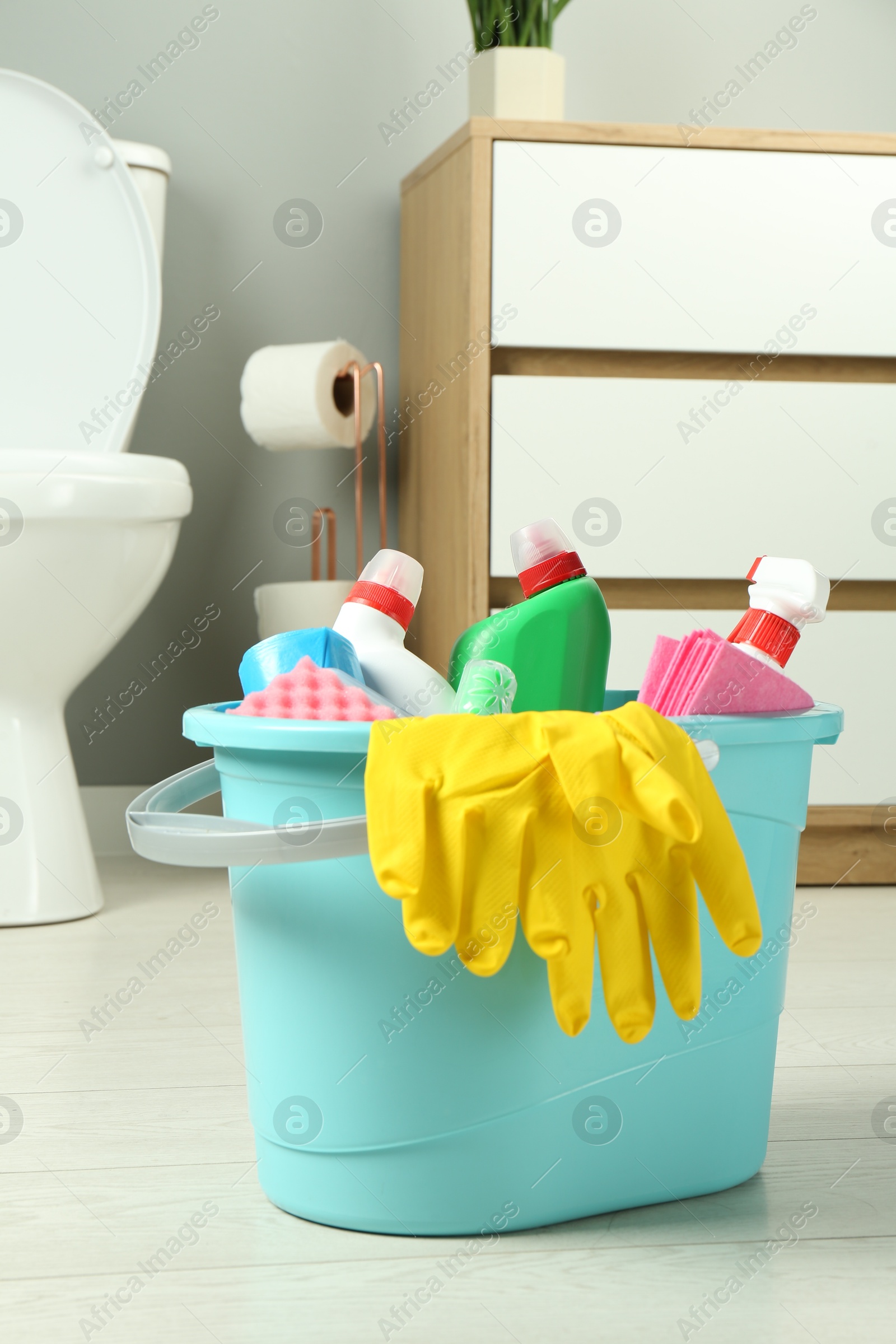 Photo of Bucket with different toilet cleaners, rag, sponge and gloves on floor in bathroom