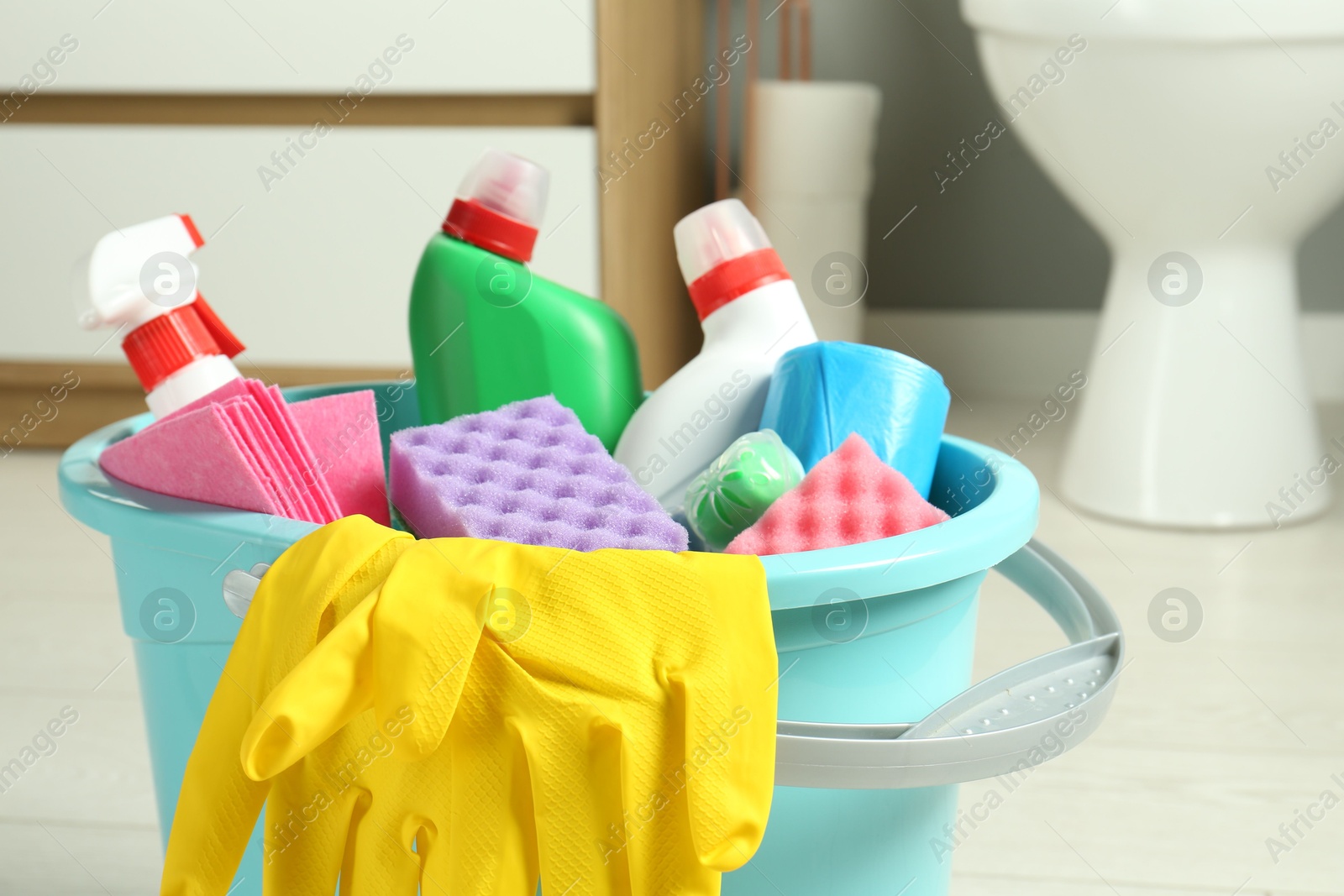 Photo of Bucket with different toilet cleaners, rag, sponge and gloves in bathroom, closeup