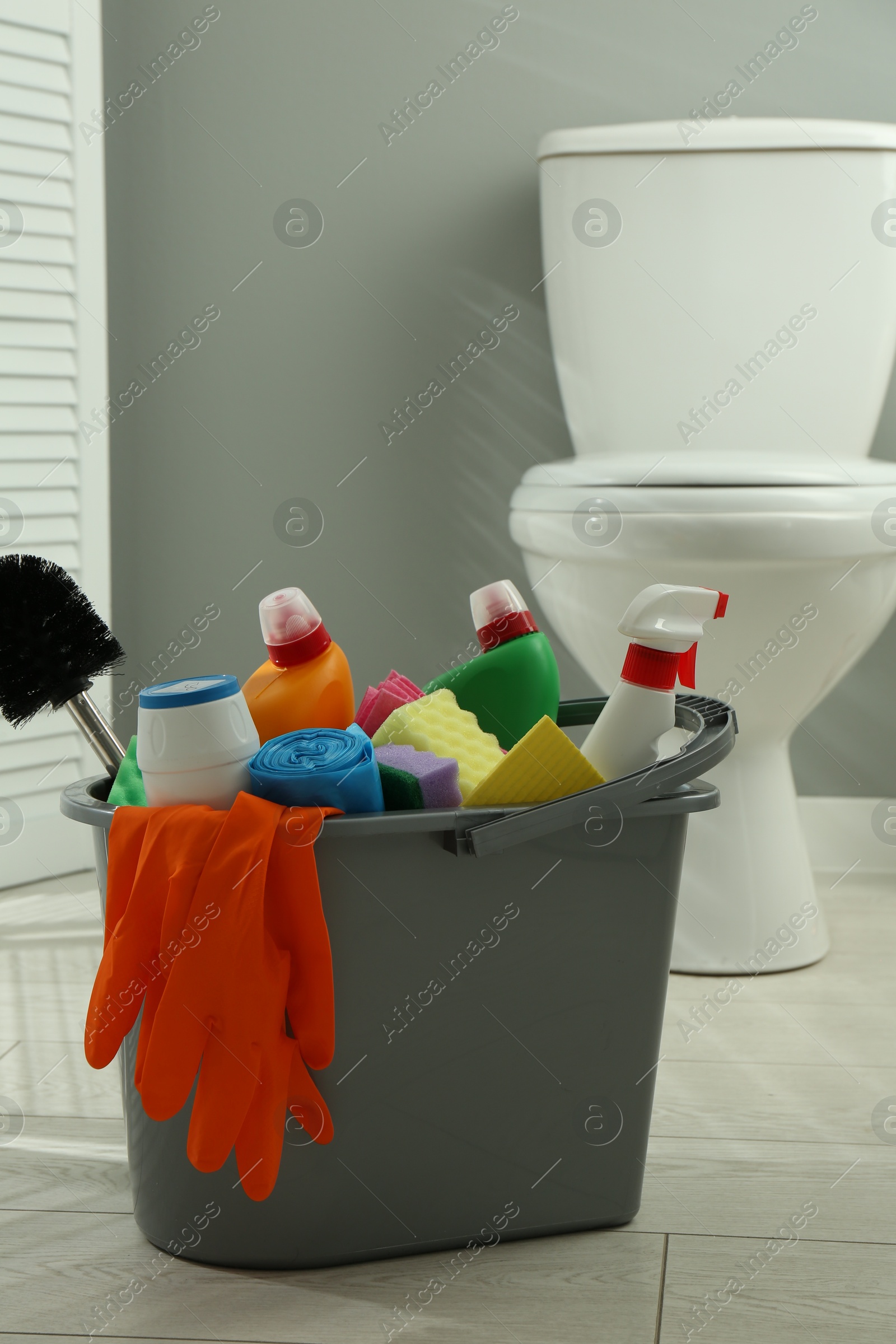 Photo of Bucket with different toilet cleaners, sponges, brush and gloves on floor in bathroom