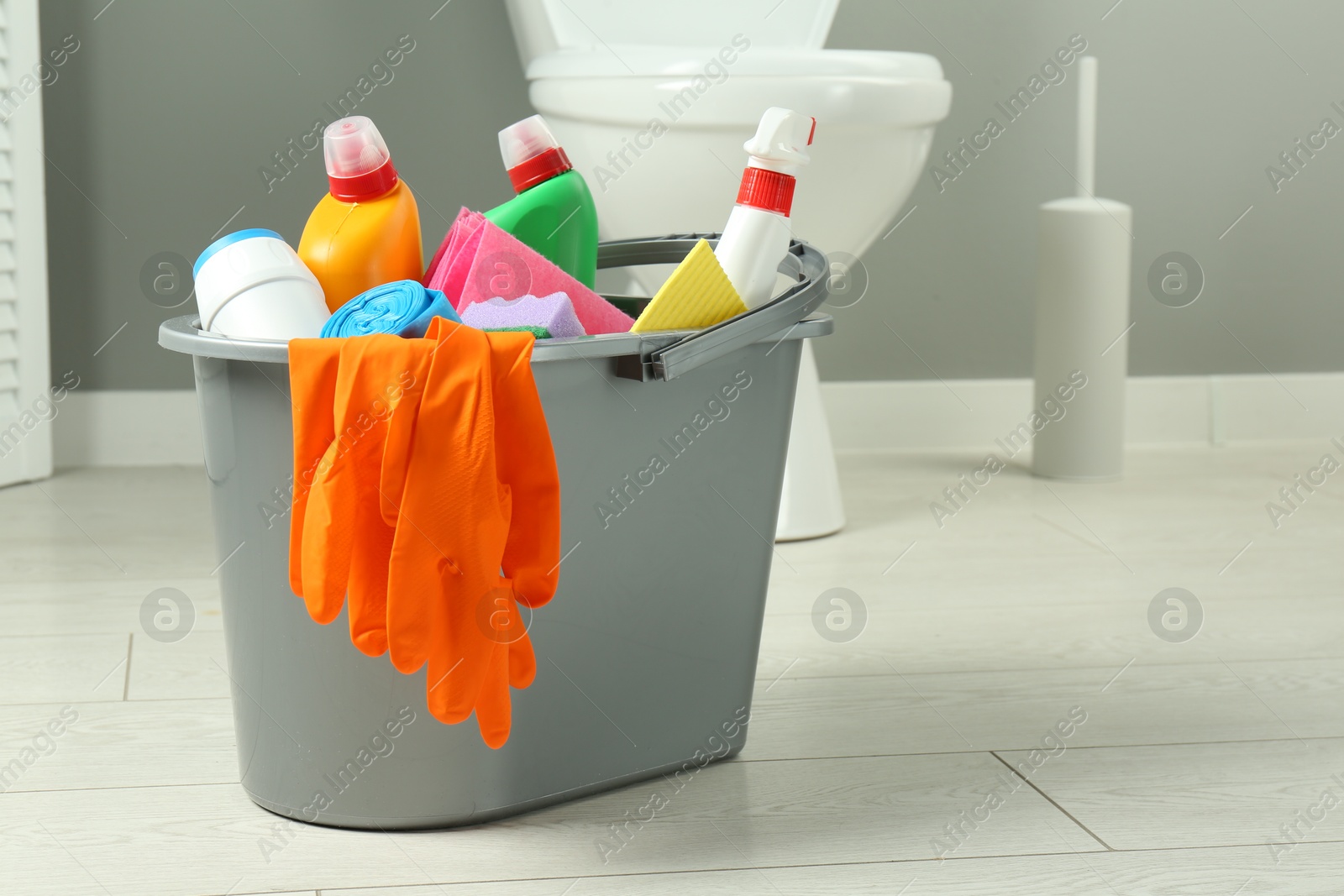 Photo of Bucket with different toilet cleaners, sponge and gloves on floor in bathroom