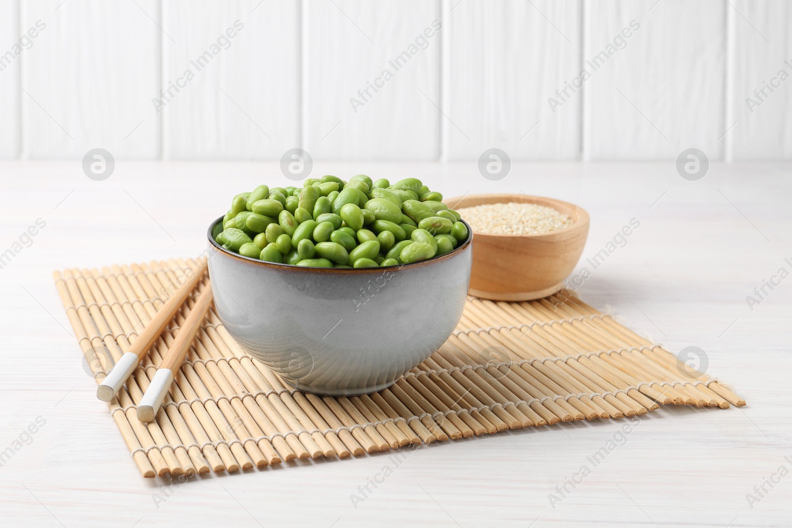 Photo of Fresh edamame soybeans in bowl, chopsticks and sesame seeds on white wooden table