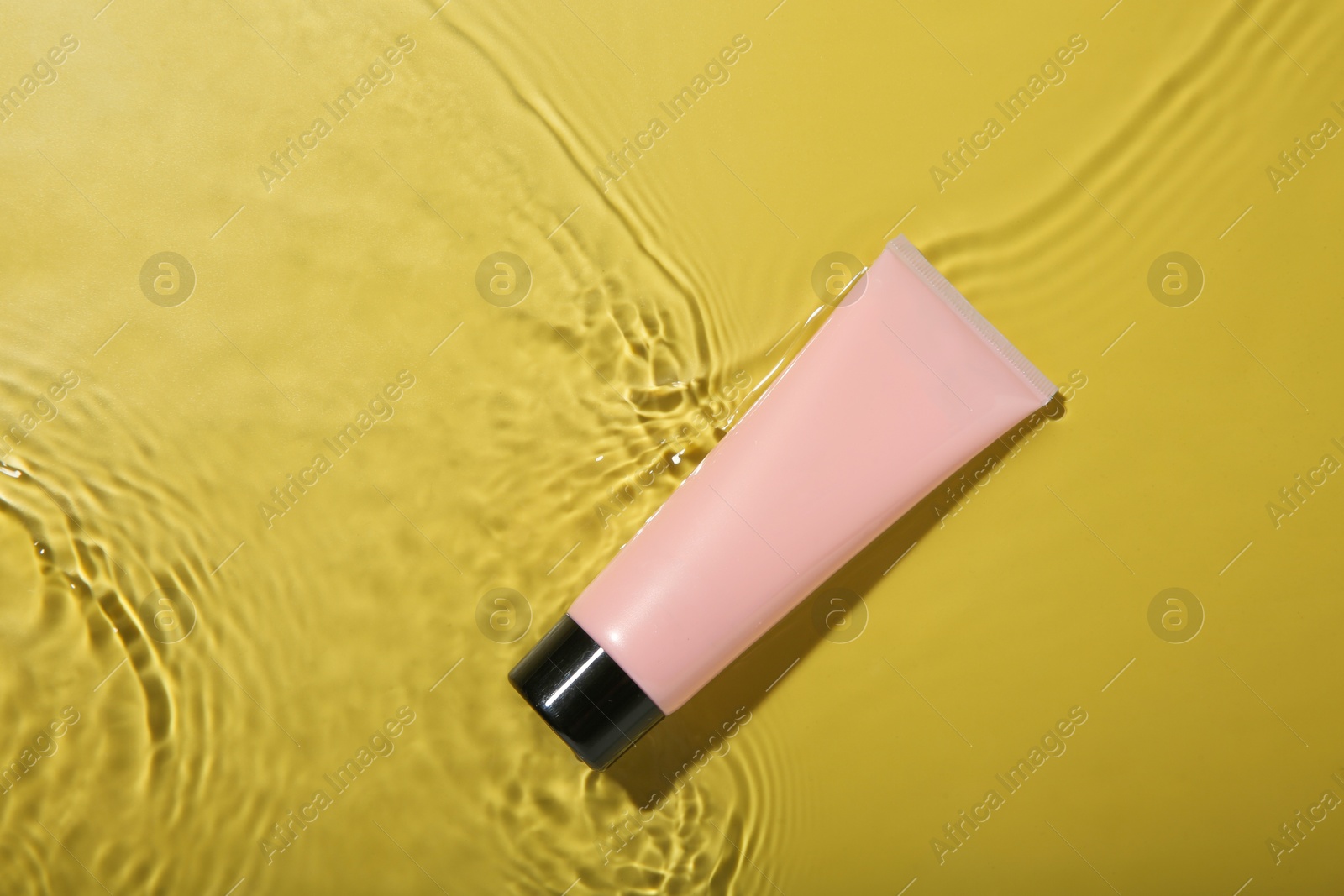 Photo of Tube with cream in water on yellow background, top view. Cosmetic product