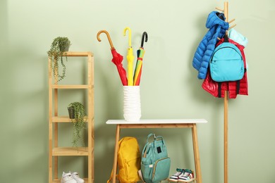 Colorful umbrellas in holder, rack with jackets, backpacks and houseplants near green wall