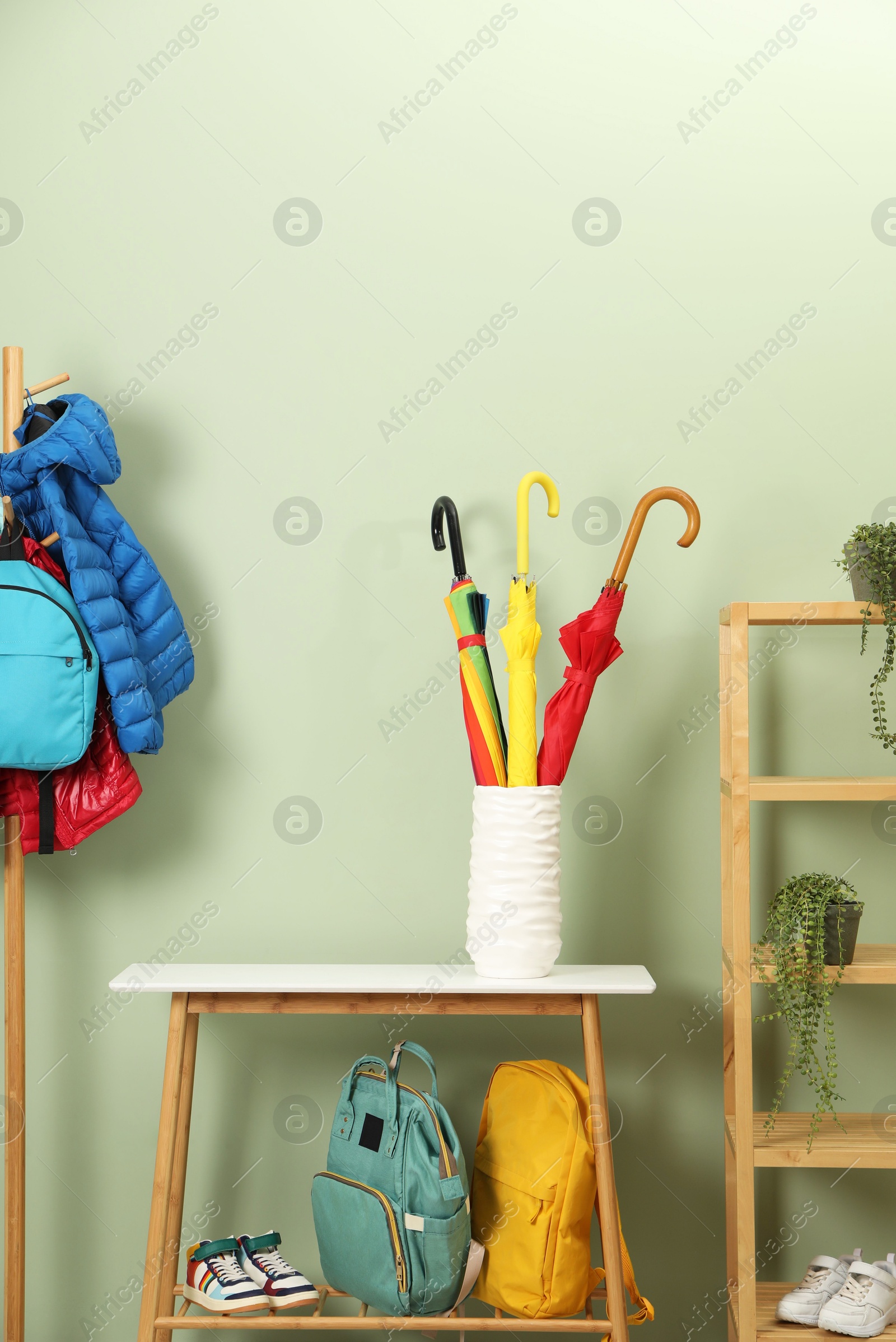 Photo of Colorful umbrellas in holder, rack with jackets, backpacks and houseplants near green wall